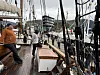 Folk går på dekket til en seilbåt i havna, med master, tauverk og flere båter i bakgrunnen.
