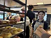 To personer står ved en bod med bakverk og brød under en innendørs festival.