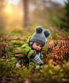 Lite barn i grønn jakke og grå lue sitter i skogbunnen blant gress og blader.