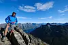 En turgåer står på en steinete fjellkant med vid utsikt mot fjell og fjord under blå himmel.