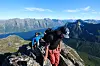 To personer går på en steinete fjellrygg med blå himmel og snødekte fjell i bakgrunnen.