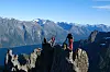 To personer står på en smal fjelltopp over en fjord omgitt av høye fjell.