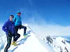 To personer står på en smal snødekt fjellrygg med fjelltopper og blå himmel.
