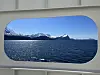 Utsikt over blå fjord med snødekte fjell i bakgrunnen, sett gjennom et avrundet vindu.
