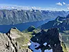 Utsikt fra en fjellrygg mot blå fjord, bratte fjell og snøflekker under en klar himmel.