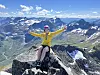 En person i gul jakke sitter på en fjelltopp med utsikt over snødekte fjell og daler.