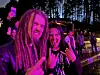 To smilende personer i svart klær viser metaltegn foran et publikum i rosa scenelys.