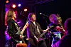 Tre musikere spiller hardrock på en сцене med fargede lys og publikum i forgrunnen.