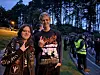 To personer i metalantrekk står foran en folkemengde utendørs ved en skogkledd arena i skumringen.