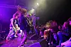 Et rockeband spiller på en scene med publikum tett foran, noen med hodene kastet frem i lys fra lilla sceneeffekter.