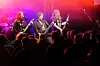 Tre musikere opptrer på en utendørsscene foran et tett publikum i rødt og lilla scenelys.