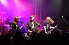 Et rockeband spiller på scene foran publikum i mørket, med lilla lys og trommesett i bakgrunnen.