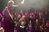 En gitarist står med ryggen til kamera foran et publikum som hever hendene under scenelys.