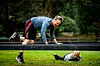 To personer på gresset i et utendørs treningsområde, én på hender og knær og én liggende under et lavt hinder.