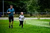 To personer løper på en grønn gressbane med trær og gjerde i bakgrunnen.