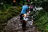 Flere personer vasser gjennom en smal, gjørmete kanal omgitt av høyt gress og vegetasjon.