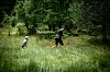To personer går eller løper i et høyt gressfelt med skog i bakgrunnen.