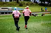 To personer løper på gresset i et uteområde med små hinder og en tilhenger i bakgrunnen.