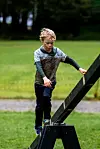 Et barn med gjørmete klær balanserer på et svart trehinder på en gresslette.