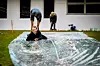 To personer arbeider med en stor plastduk fylt med vann på et gressområde foran en bygning.