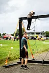To menn ved et stort svart hinder på en gressbane, med tau, kjegler og en gruppe mennesker i bakgrunnen.