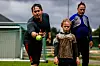 En mann i mørke klær kaster en tennisball mot en gutt på en gressbane, med en tredje person i bakgrunnen.