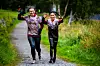 To personer løper på en grusvei mellom høyt gress og trær.