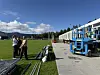 To personer ved en gressbane med utstyr og en blå løfteplattform på høyre side.