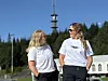 To kvinner i hvite skjorter står og snakker utendørs foran trær, et tårn og en lys bygning.