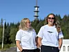 To kvinner i hvite T-skjorter smiler utendørs foran skog og et høyt tårn.