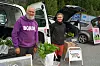 To personer står ved åpne bilbakluker med grønnsaker og varer på en utendørs markedsplass.