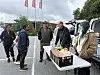 Kunder samles rundt et bord med kasser og poser ved et utendørs utleveringspunkt.