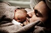 Nyfødt baby ligger tett inntil en mann under et brunt teppe.