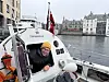 Person i gul lue lener seg ut av en liten båt i en havn med bybebyggelse i bakgrunnen.