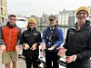 Fire menn i friluftsklær står på en brygge ved vannet og holder hendene åpne mot kameraet.
