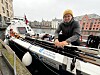 En roer i gul lue sitter i en ekspedisjonsbåt ved en kai i et bymiljø.