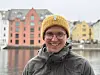Smilende mann med gul strikkelue og briller foran fargerike bygninger ved vannet.