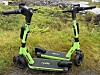 To grønne Ryde-elsparkesykler står parkert på gress og steingrunn.