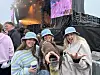 Tre smilende festivalgjengere i hatter poserer foran en konsertscene.