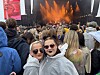 To smilende kvinner i regnponchoer og solbriller foran en konserttribune med scenelys.