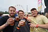 Tre menn smiler og holder ølglass foran en festivalarena med folkemengde og pariserhjul i bakgrunnen.