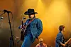 Gitarist i cowboyhatt synger på en scene med bandmedlemmer og gult scenelys i bakgrunnen.