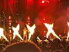 Konsertscene med flere flammer, rødt lys, artister i silhuett og publikum i forgrunnen.