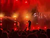 To silhuetter opptrer på en røykfylt scene foran et tett publikum i rødt lys.