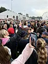 Tett folkemengde foran et stort telt, med en mobiltelefon i forgrunnen og pariserhjul i bakgrunnen.