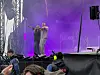 To personer står på en konsertscene med lilla lys, mens publikum ser på fra området foran scenen.