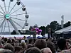 Publikum står foran en utendørsscene med artister, telt og et stort pariserhjul i bakgrunnen.