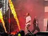 To musikere opptrer på en scene med sterke flammer, røyk og rødt lys.