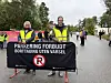 To vakter i refleksvester står bak et skilt med parkeringsforbud på en vei med mennesker i bakgrunnen.
