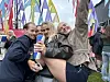 Tre personer smiler foran kameraet blant fargerike flagg på et utendørs arrangement.